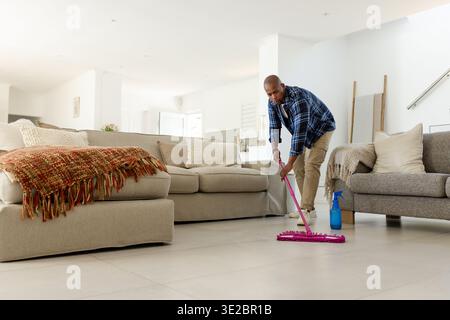 Lavare l'uomo con una camicia a quadri, spingere il mocio rosa con una bottiglia spray nel soggiorno, copiare lo spazio. Pulizia, casa, luce solare, moderno, minimalista, neutro, tessile Foto Stock