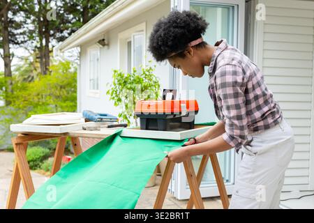 Donna afro-americana adulta in tessuto verde a quadri con cesoie su sawhorses, copia spazio. Esterno, patio, fai da te, proprietario di casa, luce naturale, forbici Foto Stock