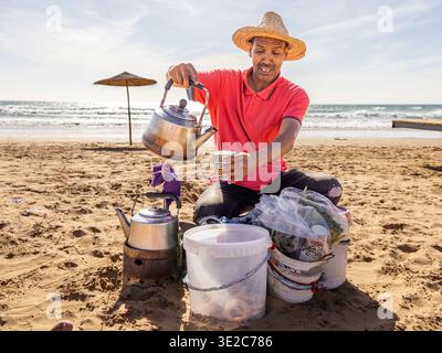 Tradizionale venditore di tè marocchino che versa tè alla menta sulla spiaggia di Taghazout, vicino ad Agadir, in Marocco Foto Stock