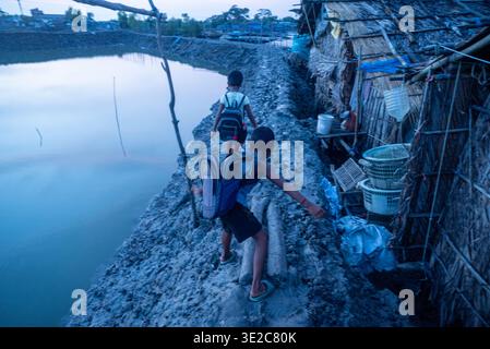 Khulna, Bangladesh - 12 luglio 2025: Veduta dei bambini che camminano lungo uno stretto sentiero fangoso tra stagni, che conduce verso capanne di paglia sotto una S rovesciata Foto Stock