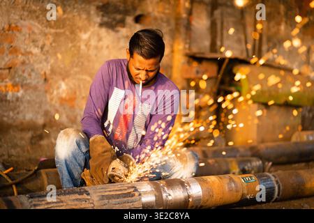 Keraniganj, Bangladesh - 11 febbraio 2023: Vista di un uomo con una sigaretta in bocca che macina metallo con scintille che volano in una scena industriale grintosa Foto Stock