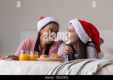 Diverse donne a letto che ridono e condividono il vassoio con occhiali e croissant OJ, indossando cappelli di Babbo Natale. Duo, accogliente, intimo, festivo, colazione, felicità, Foto Stock