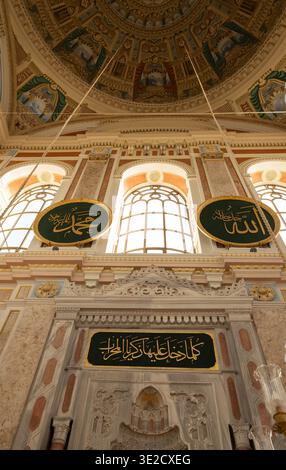 Vista ad angolo basso dell'interno di una moschea ornata con archi dorati targhe calligrafiche arabe pannelli a cupola dipinti e luminose finestre ad arco Foto Stock