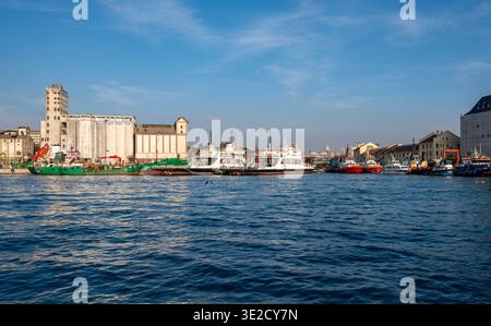 Ampio porto con barche da pesca attraccate traghetti e magazzini sul lungomare intemprati in acque blu calme sotto un cielo limpido con edifici della città Foto Stock