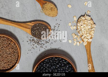 Una vista dall'alto di vari grani e legumi esposti su cucchiai di legno e in ciotole, tra cui avena, semi di chia, lenticchie e fagioli neri, contro a. Foto Stock