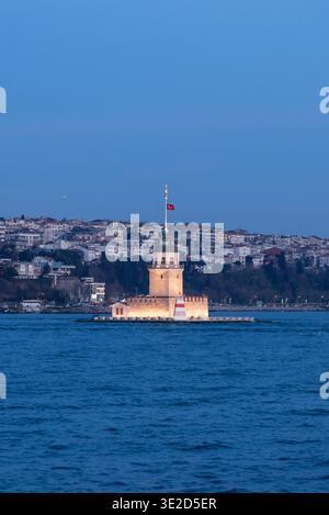 Torre della Fanciulla illuminata su un piccolo isolotto nel Bosforo al crepuscolo con bandiera turca e edifici collinari di Istanbul che creano un paesaggio di viaggio panoramico Foto Stock