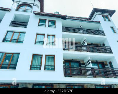 La neve copre l'area mentre cade su un edificio bianco con più balconi. La scena è ambientata in un quartiere durante una giornata invernale. Foto Stock