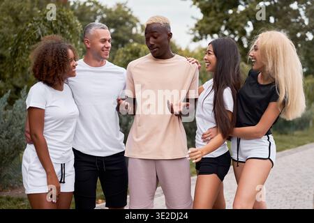 Gruppo di amici multietnici che camminano insieme nel parco estivo, ragazzo nero che racconta scherzi ad altre persone ascoltando e ridendo l'amicizia multinazionale Foto Stock