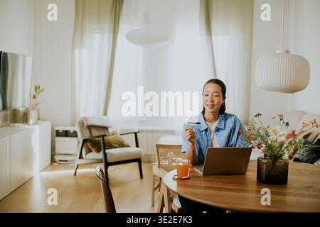 Una donna siede su un tavolo di legno con un portatile aperto davanti a lei. Sta sorridendo mentre ha una carta. La luce del sole riempie la stanza e ci sono piante Foto Stock