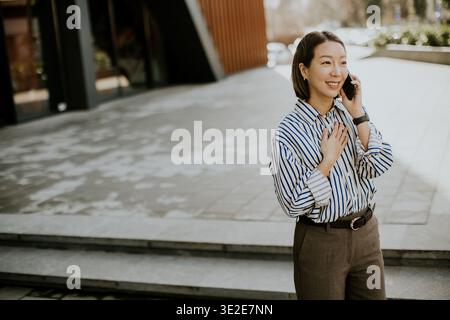 Una donna coreana si trova fuori da un edificio moderno. E' al telefono e sorride. L'impostazione sembra essere una città trafficata. E' giorno, e il sole lo e' Foto Stock