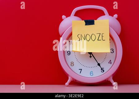 Una sveglia rosa con una nota adesiva gialla applicata sul viso con la parola scritta a mano SNOOZE. Pigrizia, concetto di procrastinazione. Foto Stock
