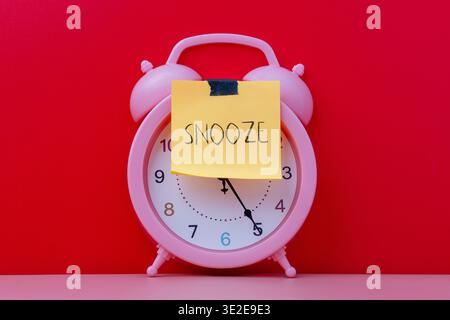 Una sveglia rosa con una nota adesiva gialla applicata sul viso con la parola scritta a mano SNOOZE. Pigrizia, concetto di procrastinazione. Foto Stock