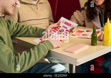 Gruppo di giovani amici seduti al tavolo che scelgono il cibo dal menu' hamburger, sorridenti e interagenti, mani e volti parzialmente visibili, gruppo multietnico che si gode un ambiente informale Foto Stock