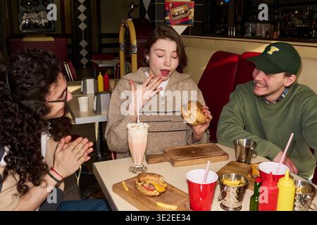 Tre giovani studenti seduti al tavolo da pranzo, donna che reagisce con sorpresa mentre tiene un hamburger, amici che sorridono e la guardano, frullato e cibo sul tavolo Foto Stock