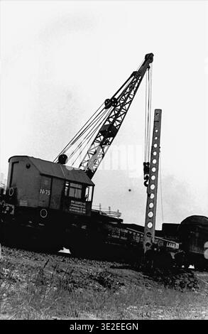 Fotografia storica in bianco e nero del 1969 che documenta una brigata di ferrovieri sovietici che installa un pesante palo in cemento lungo i binari della stazione di Slavyansk. Una gru ferroviaria MK TSUMZ-15 solleva il massiccio montante perforato mentre i lavoratori sagomati guidano manualmente la base in posizione. Questa avvincente scena archivistica mette in evidenza la combinazione di macchinari pesanti e lavoro manuale, catturando l'elettrificazione delle infrastrutture in corso e l'autentica vita lavorativa quotidiana nel tranquillo Donbass. Foto Stock