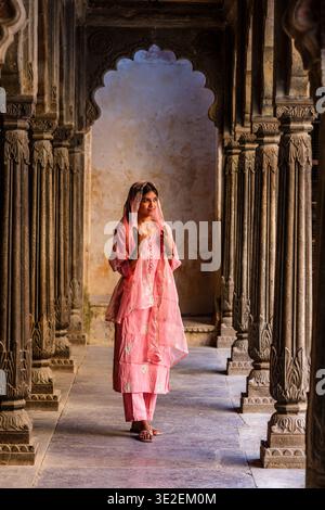 Una donna che indossa un tradizionale salwar rosa kameez si trova tra i pilastri decorati scolpiti di uno storico colonnato al Garh Palace di Bundi. Foto Stock
