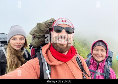 Ritratto di tre escursionisti felici in posa per un selfie su un sentiero di montagna nebbioso. L'uomo sorridente al centro indossa occhiali da sole, mentre due donne indossano beanies. Zaini visibili, con paesaggio nebbioso sullo sfondo. Foto Stock