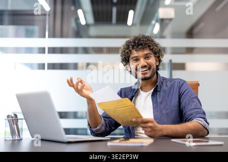Ritratto di un giovane musulmano seduto in un ufficio a una scrivania, che tiene una busta con una lettera e sorride alla macchina fotografica. Foto Stock