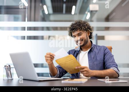 Un giovane indiano siede ad una scrivania in un ufficio e sorride mentre legge una lettera che ha ricevuto in una busta. Foto Stock