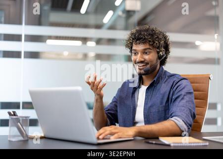 Giovane indiano che indossa la cuffia che parla in videochiamata sul laptop, gestando con le mani. Foto Stock