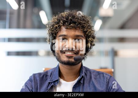 Ritratto ravvicinato di un giovane indiano in cuffia seduto in un ufficio di fronte alla fotocamera e sorridente. Foto Stock