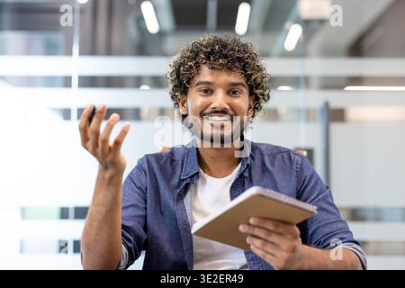 Foto ravvicinata di un giovane insegnante e formatore musulmano seduto al tavolo, tenendo una penna con un taccuino e parlando con la macchina fotografica. Foto Stock