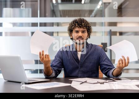 Un giovane indiano preoccupato seduto a una scrivania in ufficio, tenendo i documenti in mano e guardando la macchina fotografica con frustrazione. Foto Stock