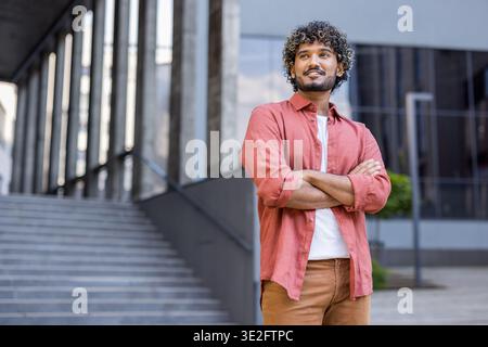 Foto di un giovane indiano sorridente in abiti informali in piedi vicino a un edificio, braccia incrociate sul petto e guardano con sicurezza di lato. Foto Stock