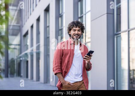 Ritratto di un giovane indiano sorridente che cammina per la città, tiene la mano in tasca, tiene il telefono nell'altra mano e guarda la macchina fotografica. Foto Stock