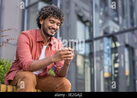 Sorridente giovane indiano seduto su una panchina all'esterno con le cuffie e con il cellulare. Foto Stock