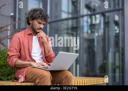 Un giovane indiano siede su una panchina vicino a un edificio di uffici e guarda attentamente lo schermo del suo notebook. Foto Stock