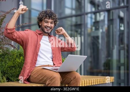 Ritratto di un uomo indiano felice in una camicia a righe seduto su una panchina all'esterno, tenendo un portatile in grembo, gioendo e alzando le braccia. Foto Stock