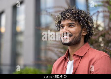 Foto ravvicinata di un giovane indiano con una camicia rossa seduto fuori e guardando di lato con un sorriso. Foto Stock