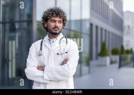 Foto ravvicinata di un serio giovane medico indiano in uniforme bianca, con uno stetoscopio in piedi con le braccia incrociate e che guarda di lato. Foto Stock