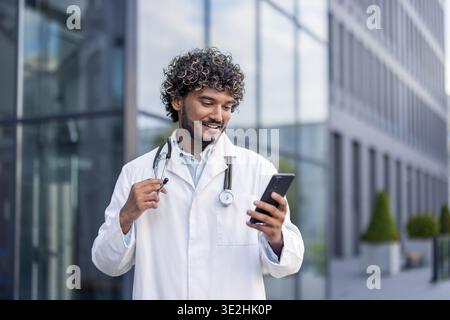 Giovane medico indiano maschile in camice bianco in piedi fuori dalla clinica e utilizzando il telefono cellulare. Foto Stock