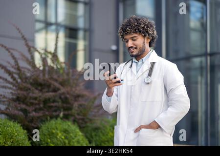 Giovane medico indiano maschio in camice bianco in piedi vicino alla clinica edificio e utilizzando il telefono cellulare. Foto Stock