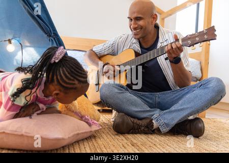 Padre e bambino afroamericano che suonano la chitarra in camera da letto, figlia che tiene il teddy. Strumento, bambino, famiglia, rustico, caldo, tenda, tappeto || modello rilasciato Foto Stock