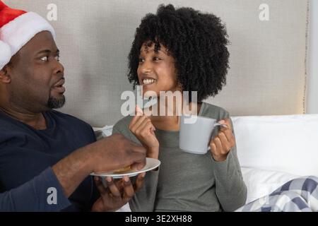 Coppia afroamericana seduta a letto tenendo tazze e piatti a costine, indossando il cappello di Babbo Natale. Accogliente, romantica, camera da letto, ambiente, piano, pigiama, ceramica || modello rilasciato Foto Stock