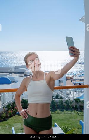 Vacanze e viaggi. Donna felice in costume da bagno che scatta selfie su un telefono cellulare in piedi sul balcone della camera d'albergo, vista mare || modello rilasciato Foto Stock