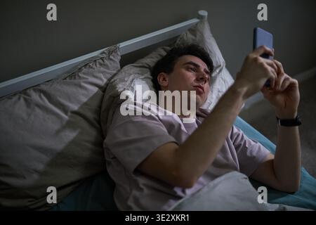 Giovane uomo caucasico adulto sdraiato a letto che regge lo smartphone sopra il viso, che mostra espressioni neutrali o contemplative mentre utilizza il dispositivo, che poggia la testa sul cuscino, che interagisce con lo schermo Foto Stock