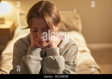 Giovane donna adulta caucasica seduta sul letto che tiene il collo e piange con l'espressione facciale angosciata, mostrando segni visibili di dolore emotivo e vulnerabilità nell'ambiente della camera da letto Foto Stock