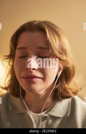 Verticale di una giovane donna caucasica adulta che chiude gli occhi con espressione emotiva, ascolta la musica attraverso auricolari cablati, mostra sottili lacrime sulle guance, trasmettendo sensazioni profonde Foto Stock