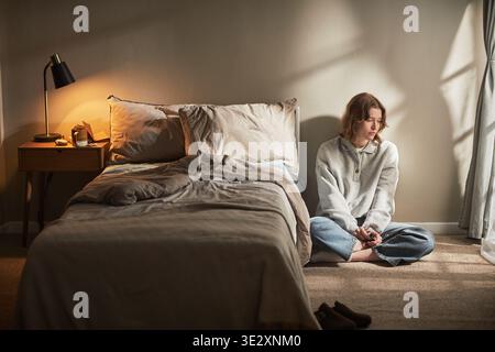 Giovane donna caucasica adulta seduta sul pavimento accanto al letto che guarda verso il basso con un'espressione premurosa, mani strette, luce del sole che scorre attraverso la finestra che proietta ombre sul muro Foto Stock