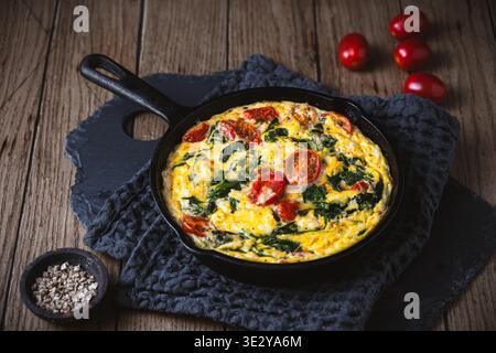 Frittata con spinaci e pomodoro in una padella di ghisa su sfondo di legno, spazio copia Foto Stock