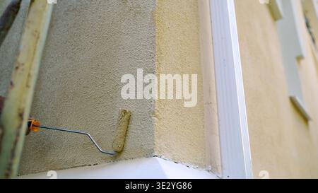 Un'immagine ad angolo basso del braccio di un pittore che applica un nuovo strato di vernice beige alla facciata in stucco testurizzato di una casa con un rullo. Quell'uomo sta lavorando Foto Stock