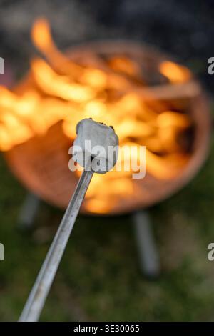 Immagine verticale del marshmallow sulla tostatura del bastone sul fuoco con fiamme luminose sullo sfondo, campeggio all'aperto e concetto di serata accogliente Foto Stock