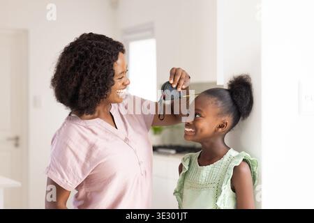 La madre di misura con la parte superiore rosa che tiene il metro sopra la ragazza in un abito verde nella cucina di casa Foto Stock