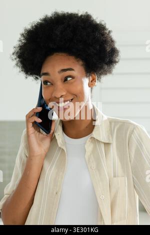 Donna afroamericana che indossa una camicia a righe beige che tiene il telefono all'orecchio destro, sorridendo in cucina Foto Stock