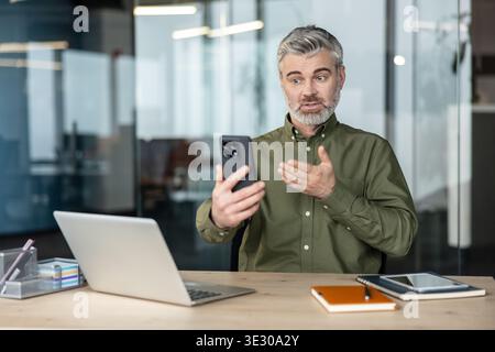 Uomo d'affari fiducioso dai capelli grigi che ha un'importante videochiamata su uno smartphone mentre è seduto alla sua scrivania in un ufficio moderno e comunica in remoto tramite la tecnologia Foto Stock