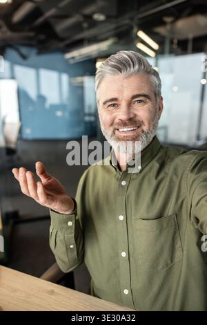 Uomo d'affari maturo con capelli grigi e barba sorridente, saluto e gesti verso la fotocamera durante una riunione virtuale o una conferenza online dal suo ufficio contemporaneo Foto Stock